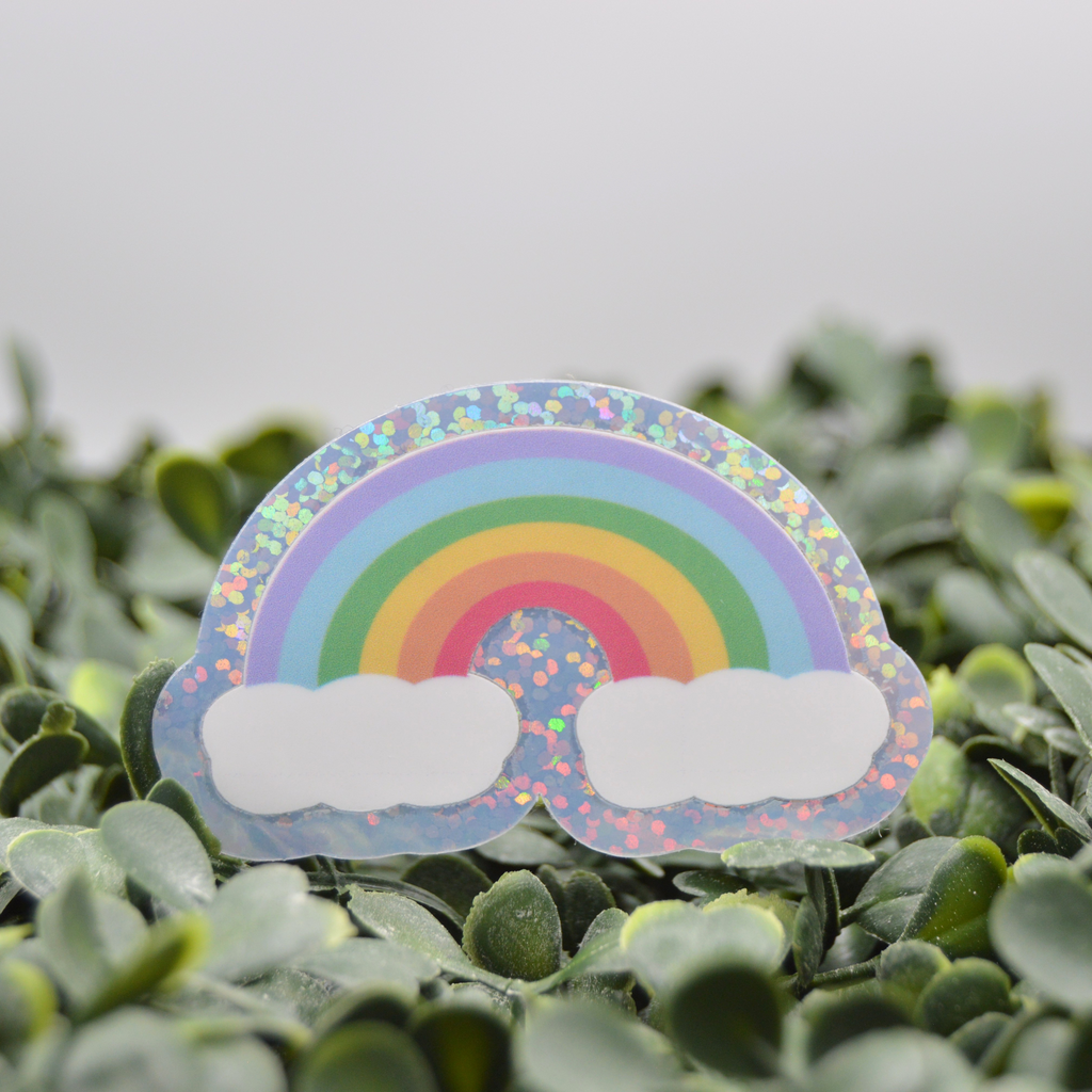 A die-cut sticker of a pastel rainbow with two white clouds, placed on a bed of green leaves. The sticker has a holographic glitter border.