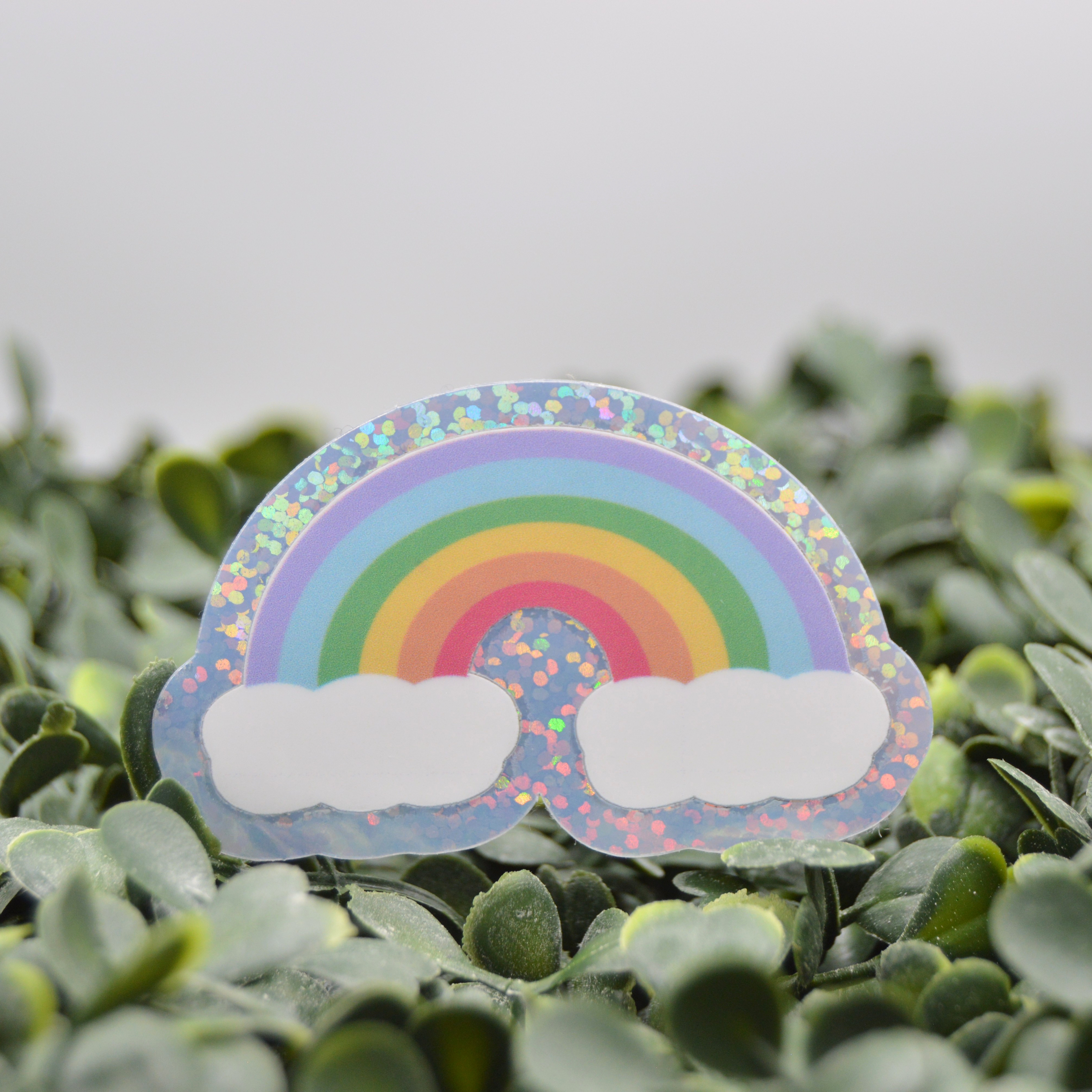 A die-cut sticker of a pastel rainbow with two white clouds, placed on a bed of green leaves. The sticker has a holographic glitter border.